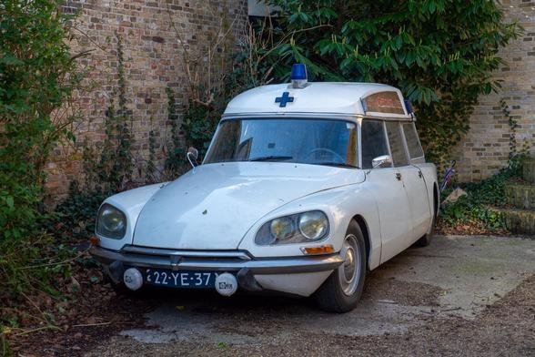 Foto eines weißen Citroën DS Autos in einem Hof, das als Krankenwagen ausgebaut ist: blaues Kreuz vorne am Dach, Blaulicht obendrauf, hinten erhöhtes Dach. Das Auto ist mit einem alten Kennzeichen versehen, weiße Schrift auf schwarzem Untergrund.