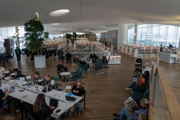 Foto mit Blick über die oberste Etage der Bibliothek: im Vordergrund Menschen an Tischen mit Leselampen und Laptops, daneben Menschen auf bequemen Stühlen Zeitung lesend. Im Hintergrund Bücherregale, weitere Tische und Stühle. Die weiße Decke ist unregelmäßig geschwungen, mit einzelnen Lichtlöchern und nur von wenigen dicken Säulen getragen. Die Außenwand ist aus Glas.