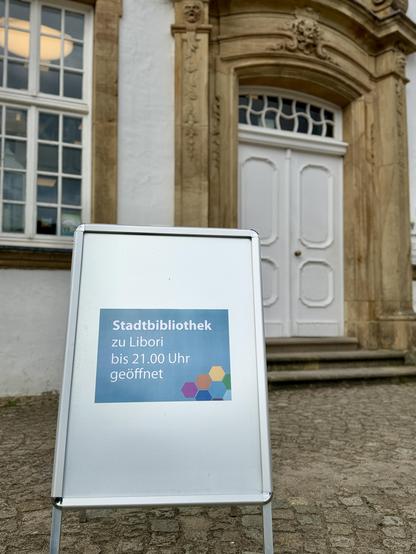 Ein Aufsteller der Stadtbibliothek Paderborn steht vor dem historischen Eingang der Bibliothek. Auf dem Schild steht: „Stadtbibliothek zu Libori bis 21.00 Uhr geöffnet“. Im Hintergrund sind die weiße Eingangstür mit barockem Sandsteinportal sowie ein Teil der Fassade zu sehen. Die Aufnahme ist leicht von unten fotografiert und stellt den Aufsteller in den Vordergrund.