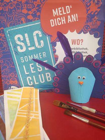 Vor einem Plakat des "Sommerleseclub" stehen Bastelvorlagen für zwei Veranstaltungen der Bibliothek: ein selbst gebastelter blauer Pfau mit Wackelaugen und Federfächer sowie mehrere bunte Lesezeichen in Gelb- Grün- und Blautönen. Daneben liegen drei Pinsel auf einem roten Untergrund. Im Hintergrund wirbt das Sommerleseclub- Plakat mit der Aufschrift "Meld dich an!"