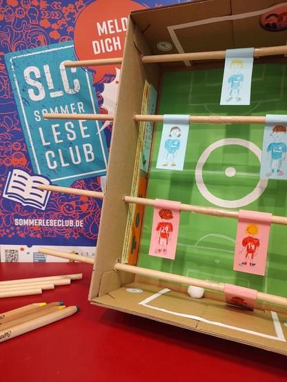 Werbung für eine Veranstaltung des Sommer Lese Clubs: Vor einem Plakat des Sommer Lese Clubs befindet sich ein aufrechter Pappkarton aus dem ein Tischkicker gebastelt wurde. Links daneben liegen Buntstifte und Strohhalme. Im Hintergrund wird ein Plakat für den Sommerleseclub mit der Aufschrift "Meld dich an!"