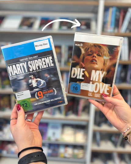 Zwei Hände halten in einer Bibliothek zwei Filme in DVD-Hüllen vor einem Regal mit weiteren Medien. Links ist „Marty Supreme“ mit Timothée Chalamet zu sehen, rechts „Die My Love“ mit Jennifer Lawrence und Robert Pattinson. Ein weißer, gebogener Pfeil zeigt von links auf den rechten Film.