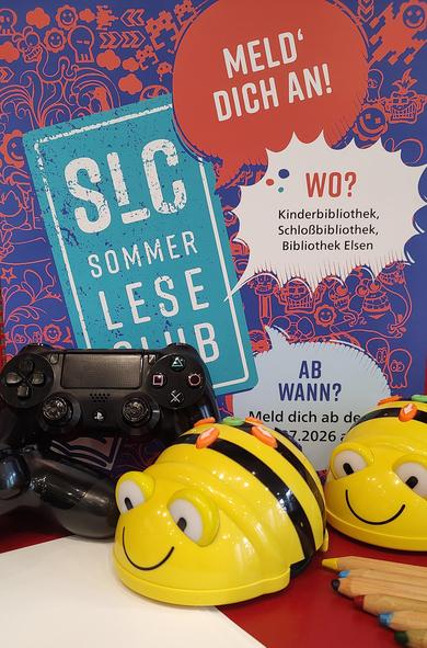Das Bild bewirbt Veranstaltungen des Sommerleseclubs. Im Vordergrung befinden sich zwei Bee-Bot Roboter, sowie zwei Playstation 4 Controller. Davor liegen mehrere Buntstifte und Papier. Im Hintergrund hängt das Sommerleseclub Plakat. Es hat den Aufdruck "Meld dich an" und die Information, dass der Sommerleseclub in der Kinderbibliothek, der Schloßbibliothek und in der Bibliothek Elsen stattfindet.