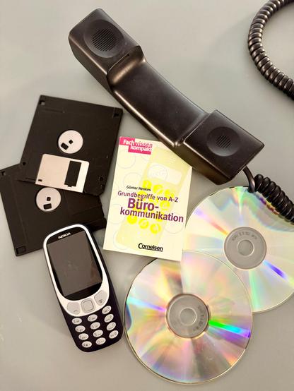 Auf einem Tisch liegen verschiedene Büro- und Kommunikationstechnik aus vergangenen Jahrzehnten: ein schwarzer Telefonhörer mit Spiralkabel, mehrere 3,5-Zoll-Disketten, ein älteres Nokia-Handy sowie zwei CDs. Daneben liegt das Buch „Grundbegriffe von A–Z Büro-Kommunikation“ von Günter Henkes aus dem Jahr 1998.