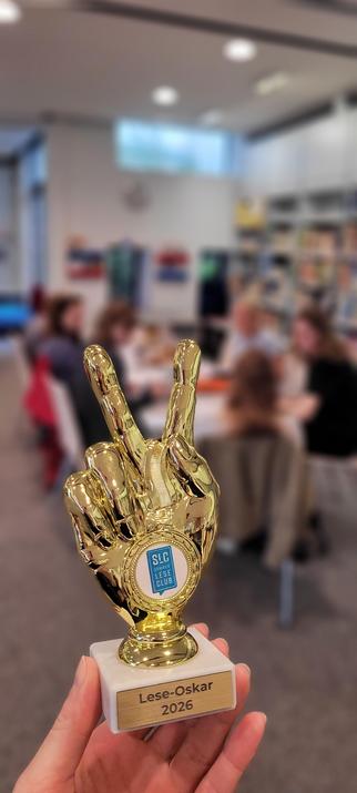 Im Vordergrund ist ein goldener Pokal in der Form einer Hand, die ein Peace-Zeichen formt. Auf dem Sockel steht Lese-Oskar 2026, auf der Hand steht Sommerleseclub. Im Hintergrund sitzen mehrere Leute an einem Tisch in der Kinderbibliothek.